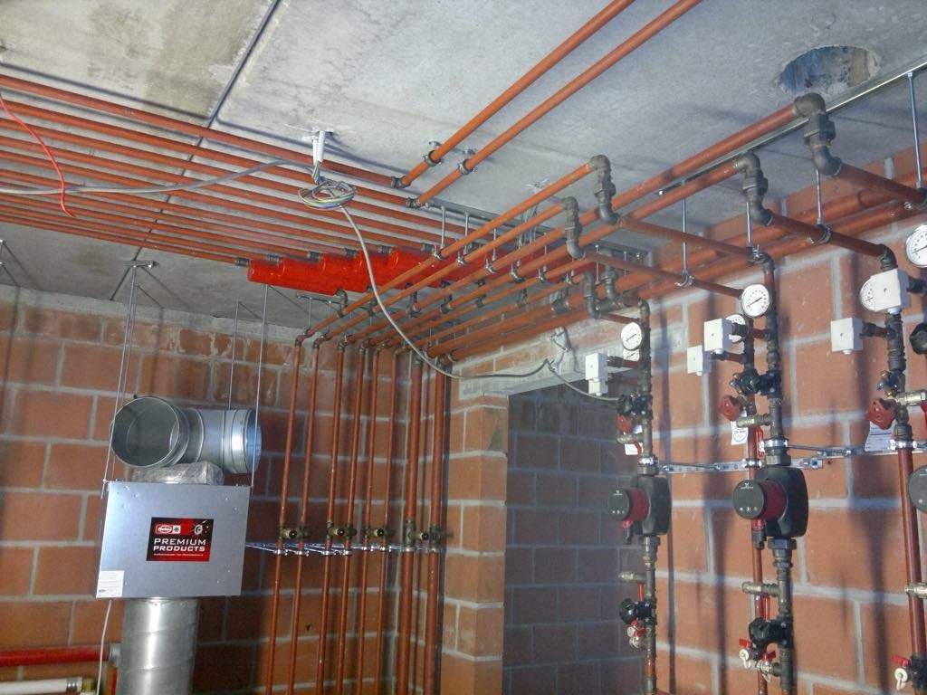 Rozbudowana instalacja rurowa z licznymi zaworami, manometrami i pompami obiegowymi w pomieszczeniu z ceglanymi ścianami i betonowym sufitem.