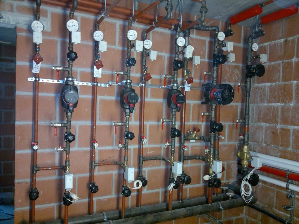 Różnorodne rury, zawory i manometry tworzące instalację grzewczą na tle ceglanej ściany, widoczne pompy obiegowe i elementy sterujące.