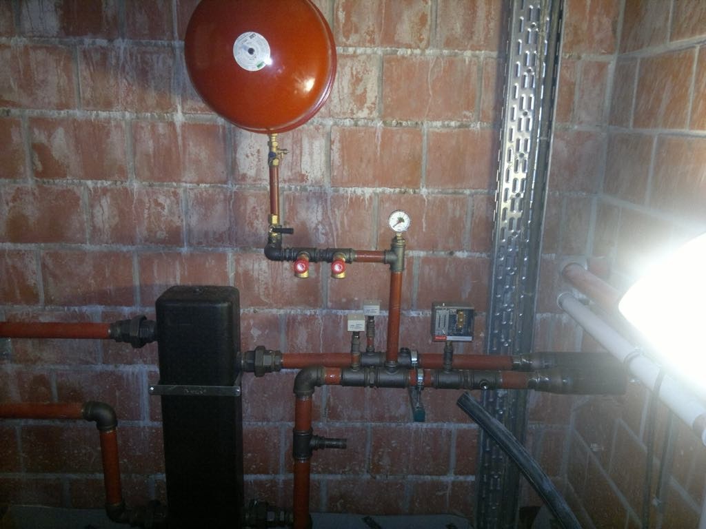 Instalacja rur miedzianych i zaworów z manometrem, zbiornik wyrównawczy na tle ściany z cegły, widoczne elementy instalacji grzewczej w piwnicy.
