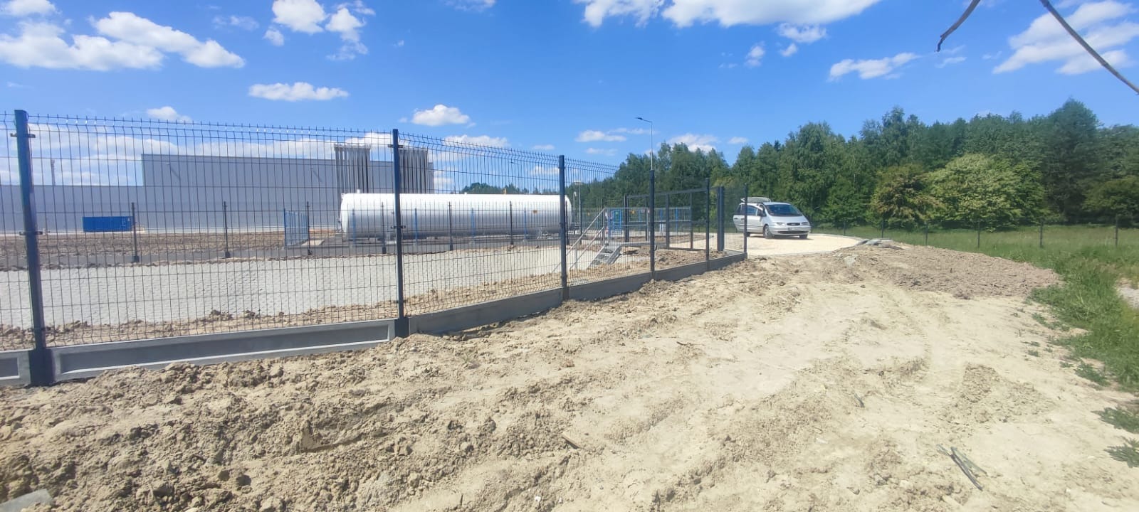 Nowe ogrodzenie panelowe z podmurówką betonową wokół przemysłowego zbiornika, widoczny srebrny samochód dostawczy na tle drzew i błękitnego nieba.