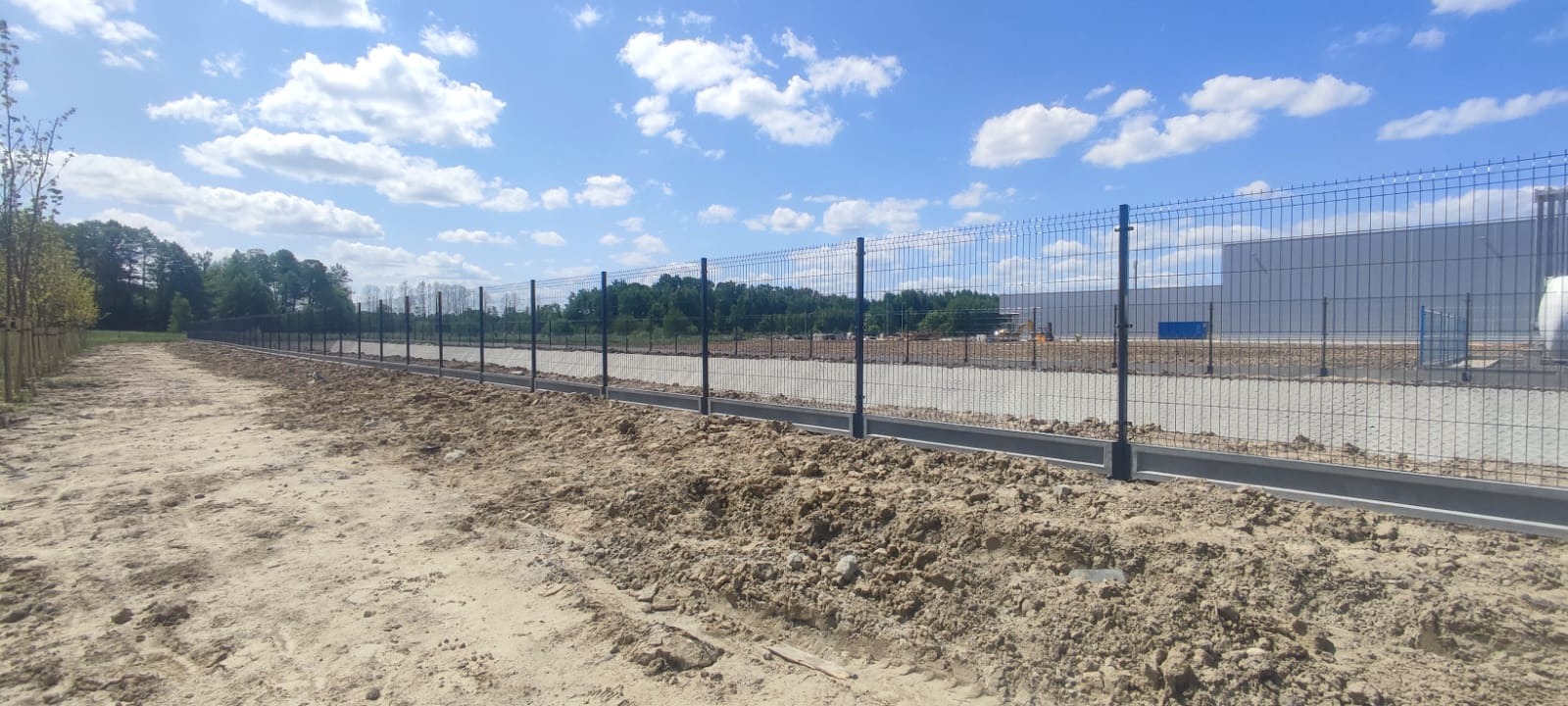 Długa, nowa siatka ogrodzeniowa z podmurówką betonową, biegnąca wzdłuż piaszczystej drogi i młodych drzew, z budynkiem przemysłowym w tle pod błękitnym niebem z chmurami.