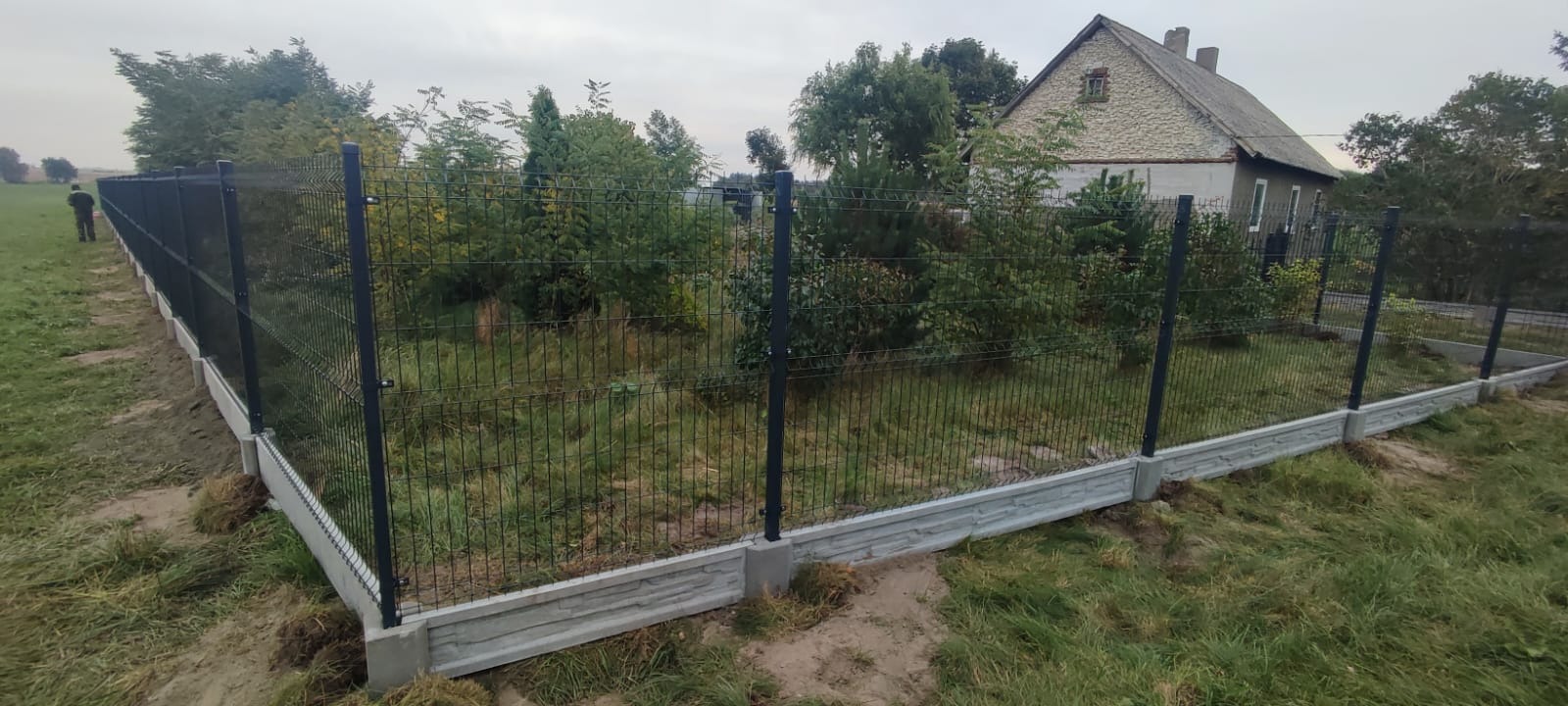 Nowoczesne ogrodzenie z siatki z podmurówką betonową, zabezpieczające posesję z widocznym domem w tle, teren nierówny, trawiasty.