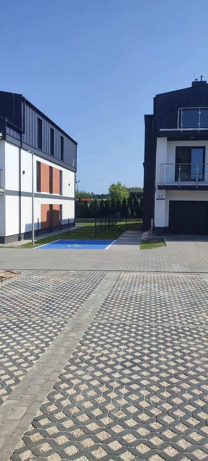 Nowoczesna posesja z szarą kostką ażurową wypełnioną drobnym kruszywem, wyznaczonym miejscem parkingowym dla osoby niepełnosprawnej i niskim, czarnym ogrodzeniem panelowym.