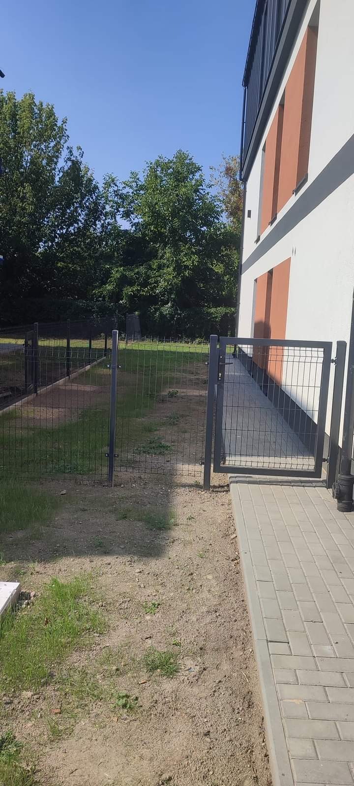 Nowoczesne ogrodzenie panelowe w kolorze grafitowym z furtką, zamontowane przy budynku z białymi ścianami i ceglastymi wstawkami, widok z zewnątrz na trawnik z fragmentami ziemi.