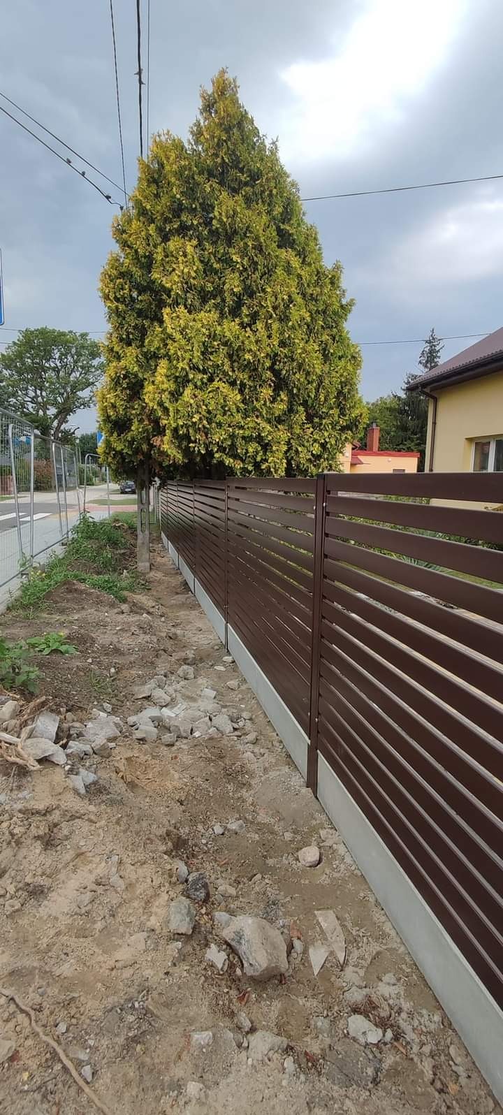 Nowoczesne, poziome ogrodzenie panelowe w kolorze brązowym z betonową podmurówką, widoczne na tle drzewa i domu jednorodzinnego.