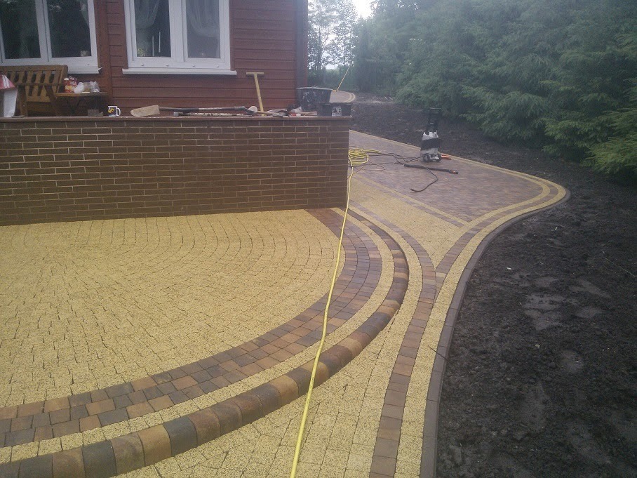 Nowo ułożony podjazd z kostki brukowej w odcieniach beżu i brązu, z widocznym wzorem łuków i narzędziami w tle, częściowo jeszcze na etapie prac wykończeniowych.