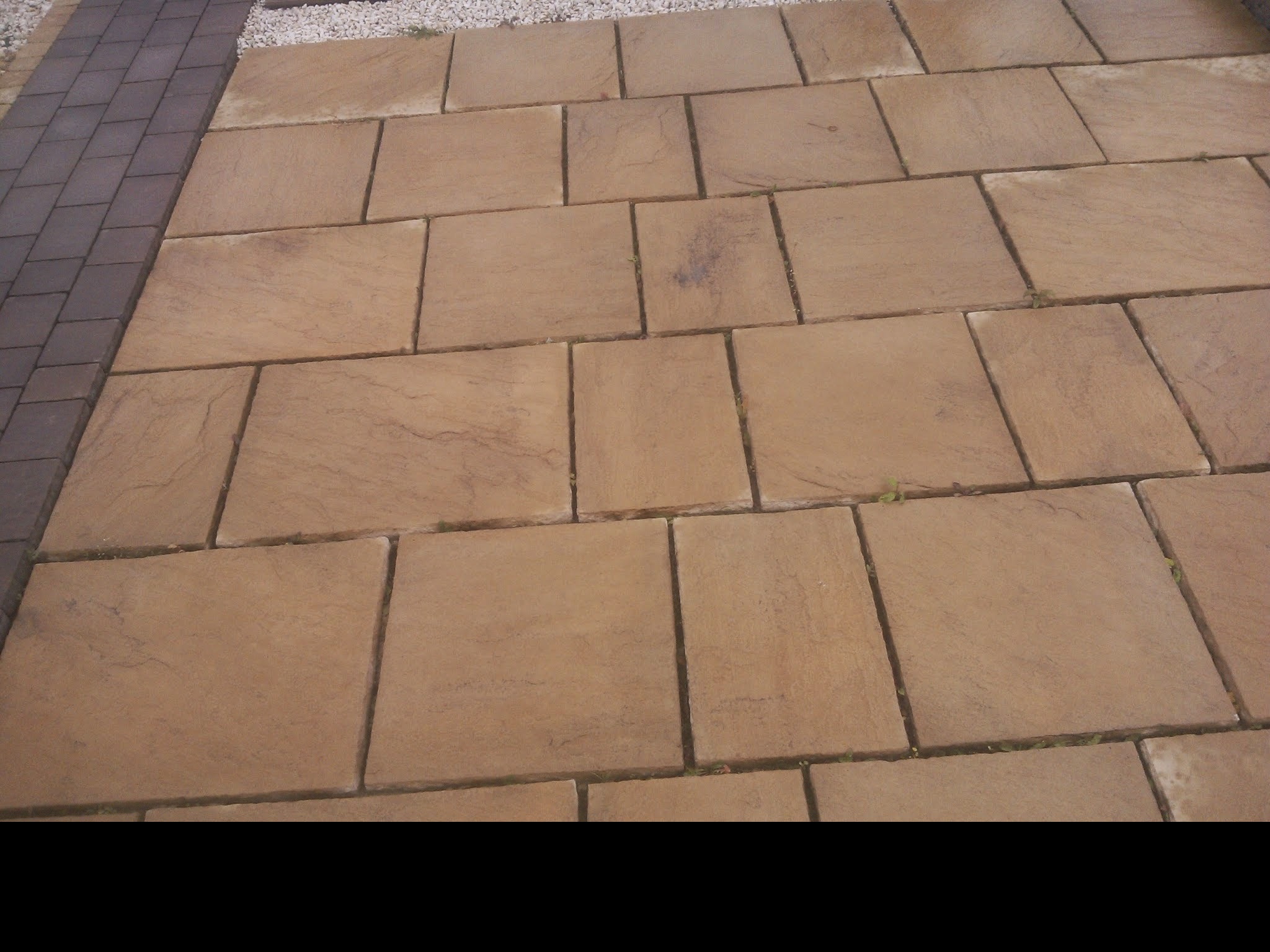 Ułożona, prostokątna kostka brukowa w odcieniach beżu i brązu, z widocznymi drobnymi roślinami wyrastającymi spomiędzy fug, obok wąski pas ciemnej kostki i jasny żwir.