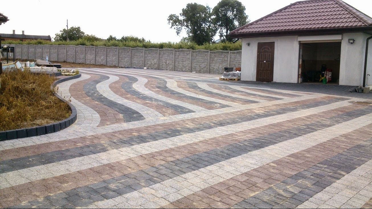 Ułożona kostka brukowa w geometryczny wzór z falującymi pasami w odcieniach szarości, brązu i beżu, prowadząca do garażu i betonowego ogrodzenia.