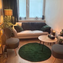 Coaching MPC - Agnieszka Kondysz - Przytulny gabinet z beżową sofą, zielonym dywanem i dekoracyjnymi lampami, oświetlony ciepłym światłem, z widoczną roślinnością i minimalistycznym obrazem na ścianie.