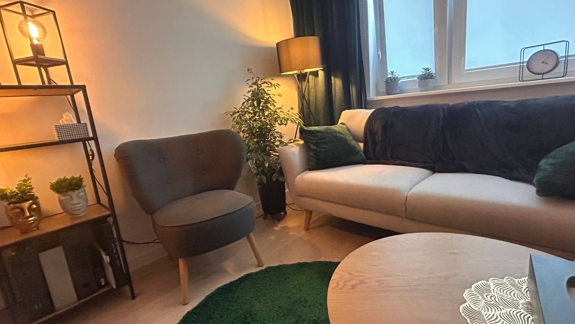 Przytulny pokój z szarym fotelem, beżową sofą z ciemnozielonymi poduszkami i pledem, okrągłym stolikiem kawowym, regałem z dekoracjami, rośliną doniczkową i lampą podłogową, tworzący relaksującą...