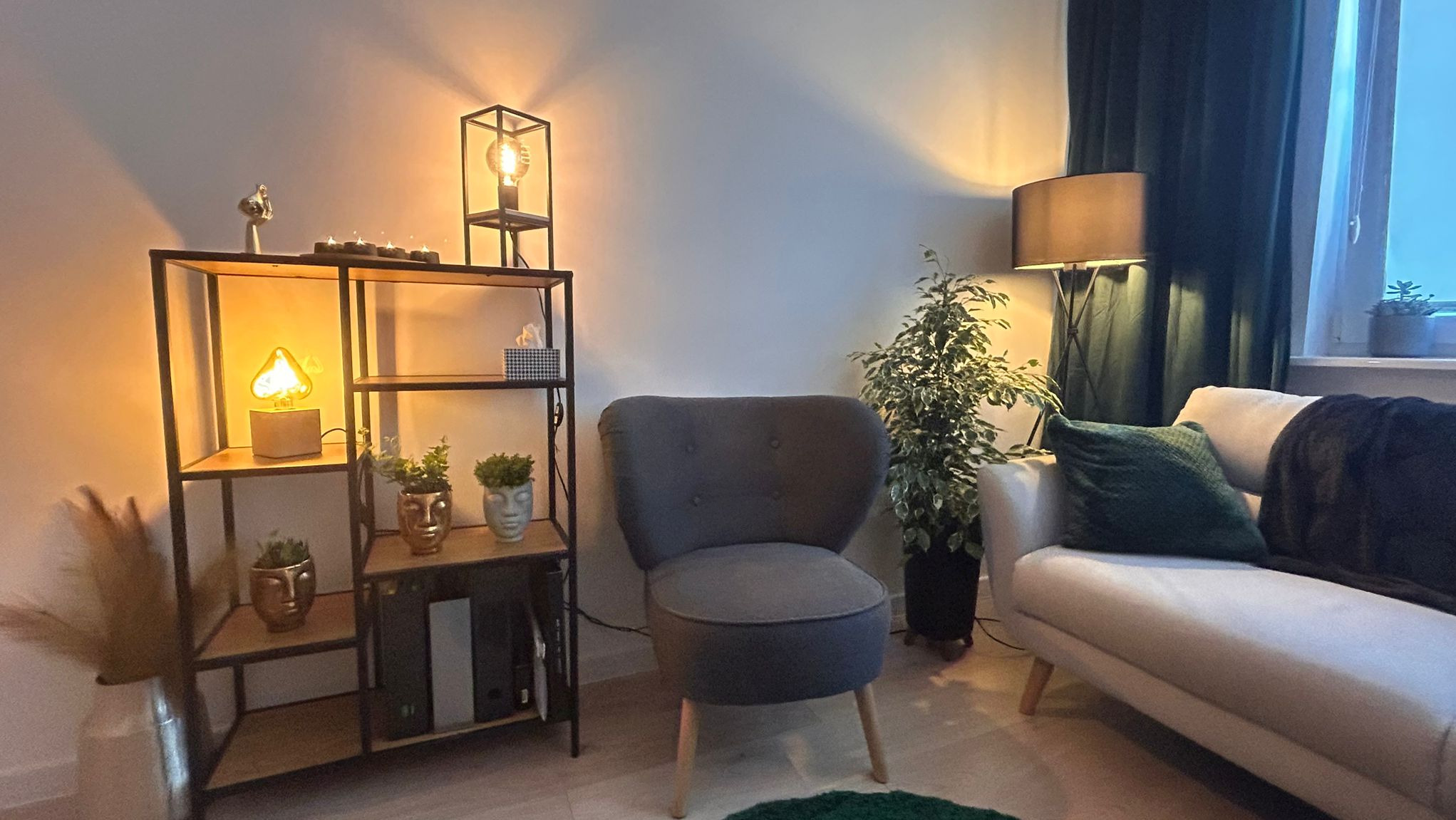 Przytulny pokój z szarą tapicerowaną pufą, regałem z dekoracjami i roślinami w doniczkach o kształcie twarzy, oraz sofą z zieloną poduszką i ciemnym pledem, oświetlony ciepłym światłem lamp.