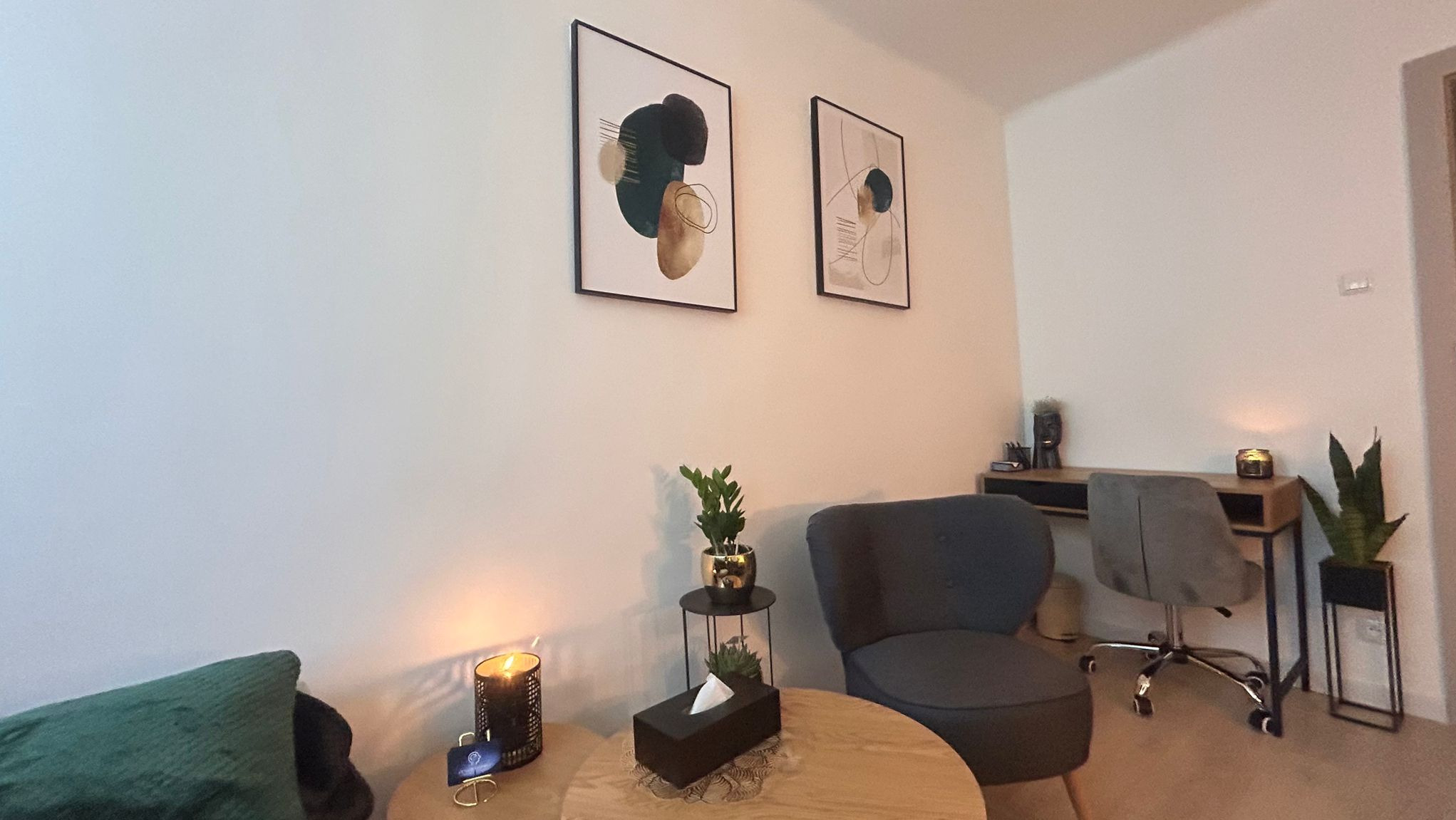 Jasne wnętrze gabinetu z dwoma abstrakcyjnymi obrazami na ścianie, okrągłym stolikiem z czarnym pudełkiem na chusteczki i zapaloną świecą, szarym fotelem oraz biurkiem z dekoracjami i rośliną...