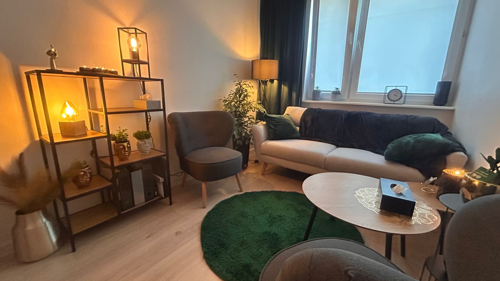 Przytulny gabinet psychoterapeuty z beżową sofą, okrągłym zielonym dywanem, metalowym regałem z dekoracjami i ciepłym oświetleniem. Na stoliku pudełko z chusteczkami.