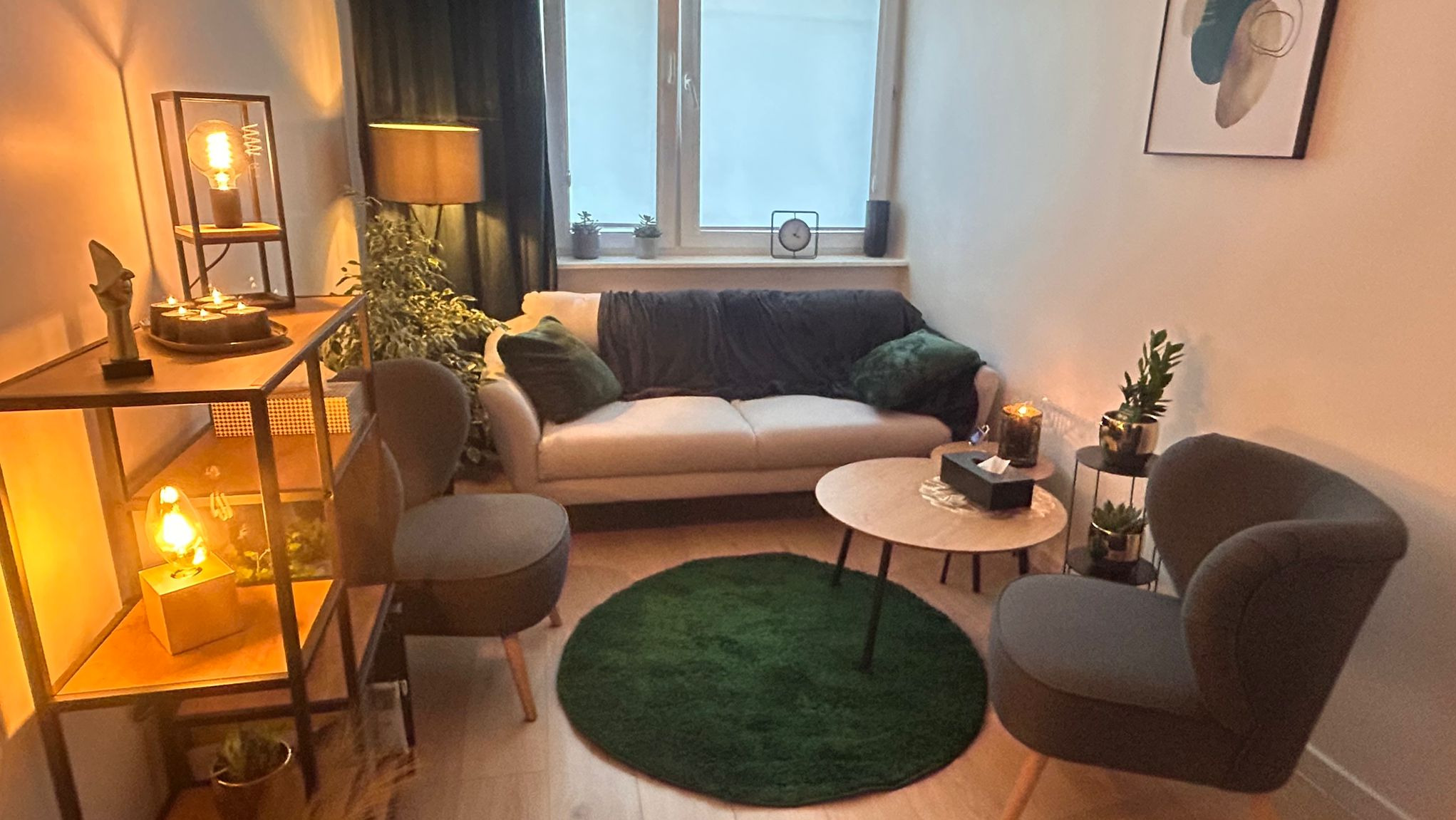 Przytulny gabinet z beżową sofą, zielonym dywanem i dekoracyjnymi lampami, oświetlony ciepłym światłem, z widoczną roślinnością i minimalistycznym obrazem na ścianie.