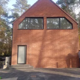 Nowoczesny dom z cegły klinkierowej z trapezoidalnymi oknami i szklanymi drzwiami wejściowymi, otoczony drzewami, z widocznymi elementami placu budowy.