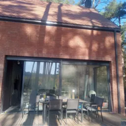 Nowoczesny dom z czerwonej cegły z dużym przesuwnym oknem, widoczne narzędzia i materiały budowlane wewnątrz, stół z krzesłami na zewnątrz, sugerujący prace wykończeniowe.