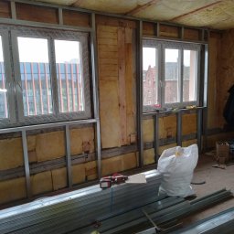 Anbud - Wnętrze w trakcie prac wykończeniowych: widoczne metalowe profile, częściowo ułożona izolacja z wełny mineralnej między drewnianymi belkami, nowe okna PCV, narzędzia i materiały budowlane na podłodze.