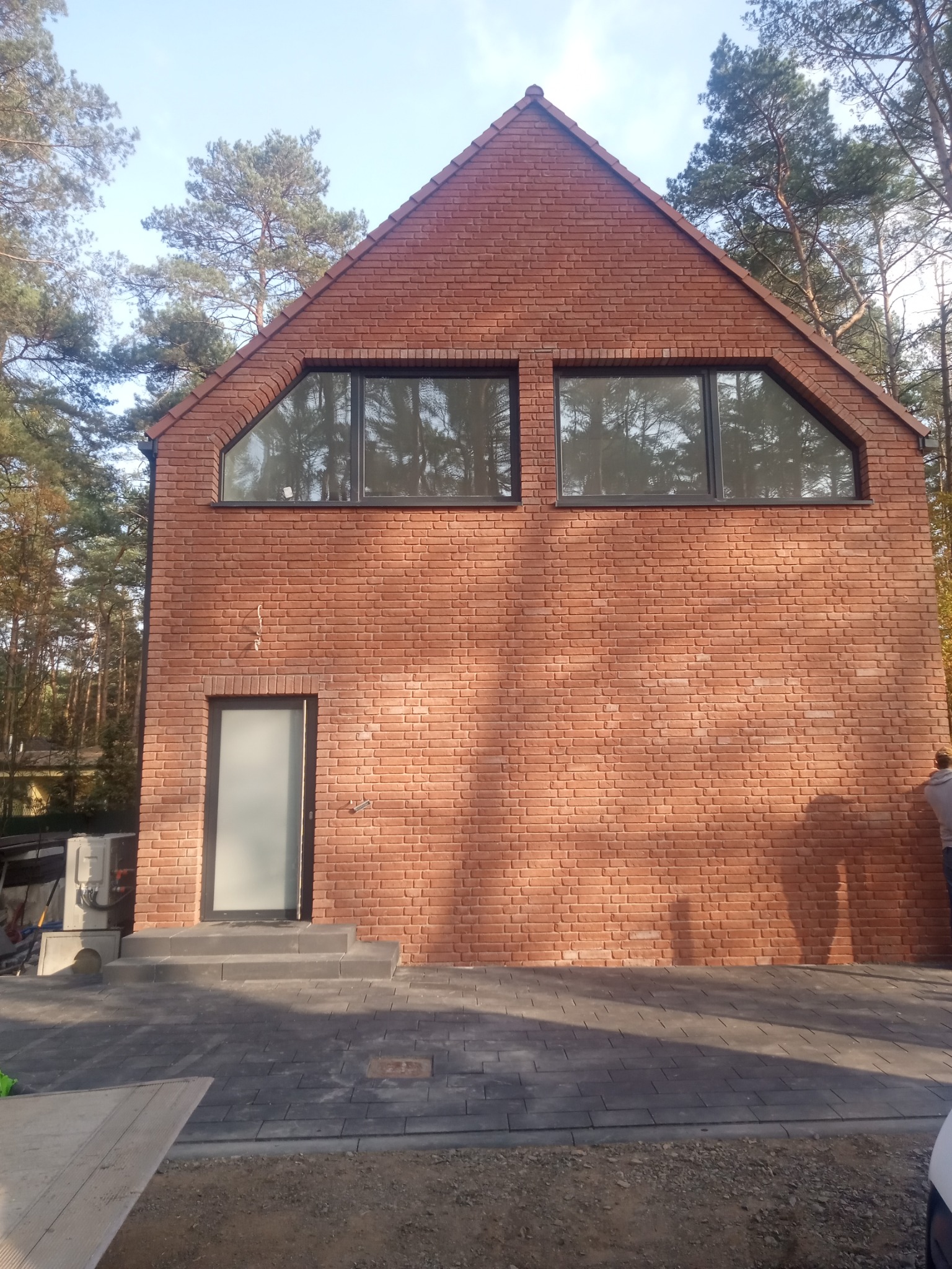 Nowoczesny dom z cegły klinkierowej z trapezoidalnymi oknami i szklanymi drzwiami wejściowymi, otoczony drzewami, z widocznymi elementami placu budowy.