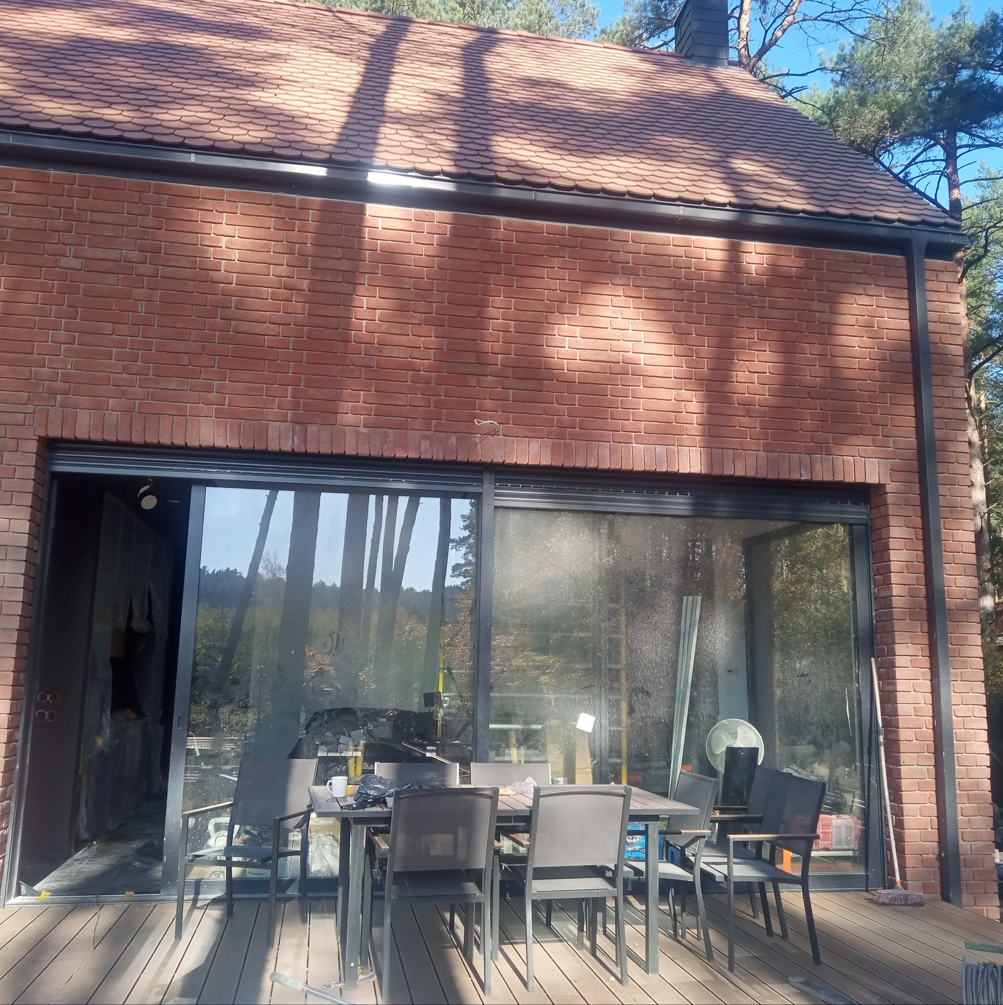 Nowoczesny dom z czerwonej cegły z dużym przesuwnym oknem, widoczne narzędzia i materiały budowlane wewnątrz, stół z krzesłami na zewnątrz, sugerujący prace wykończeniowe.