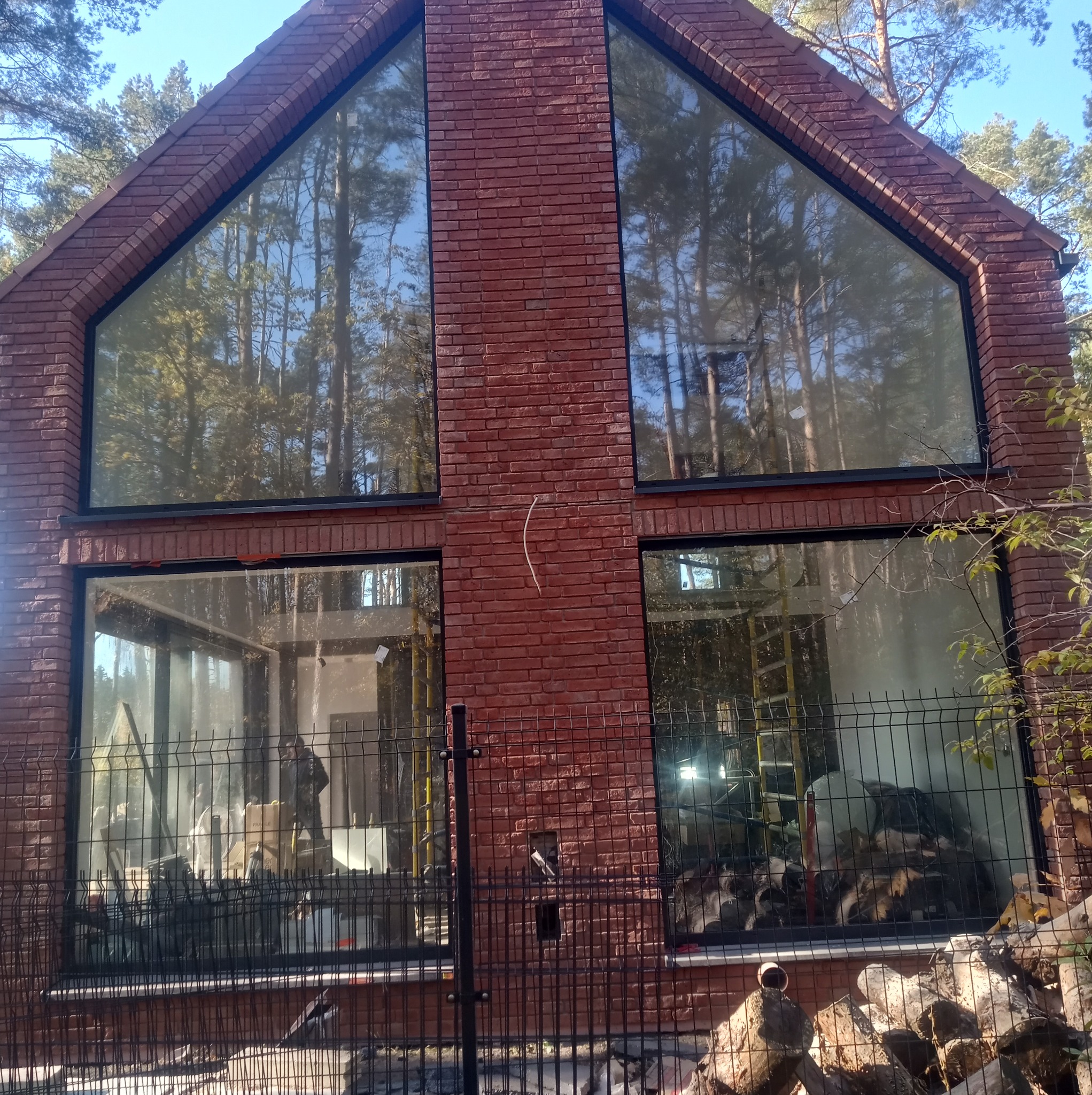 Nowoczesny dom z czerwonej cegły z dużymi oknami, widok z zewnątrz przez ogrodzenie, wnętrze w trakcie wykańczania.