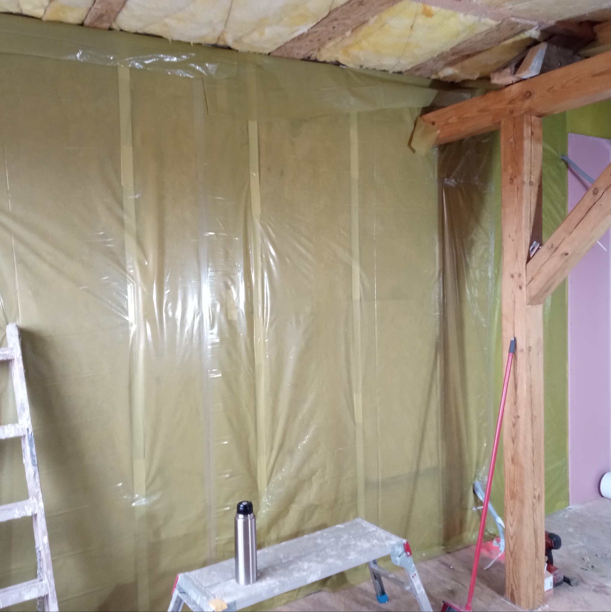Wnętrze w trakcie prac, ściana z izolacją pokryta folią, widoczne drewniane belki stropowe i aluminiowy termos na rusztowaniu.