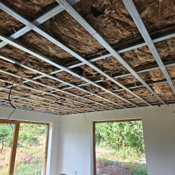 DENBLU Usługi Remontowo-Budowlane - Sufit podwieszany z izolacją z wełny mineralnej, widok z dołu na metalową konstrukcję. W tle okno z widokiem na zieleń. Częściowo widoczna sylwetka pracownika.