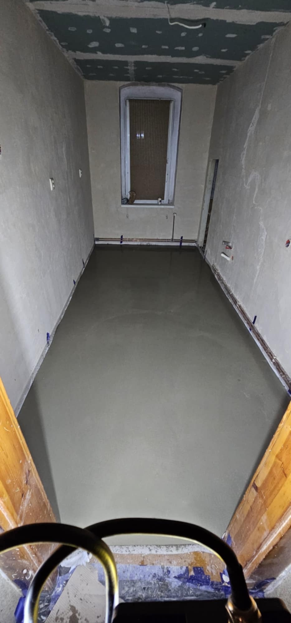 Wnętrze pokoju w trakcie wylewania posadzki, widoczne ściany po szpachlowaniu i okno z żaluzjami, niebieska taśma malarska zabezpiecza krawędzie.