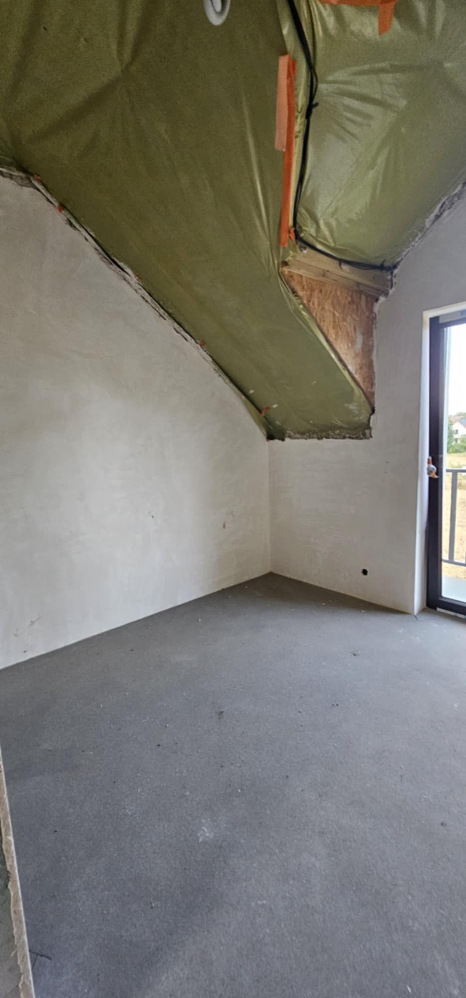 Pokój na poddaszu w trakcie prac wykończeniowych: surowe ściany po szpachlowaniu, zabezpieczona folią izolacyjną skośna część dachu, w tle widoczne okno balkonowe.