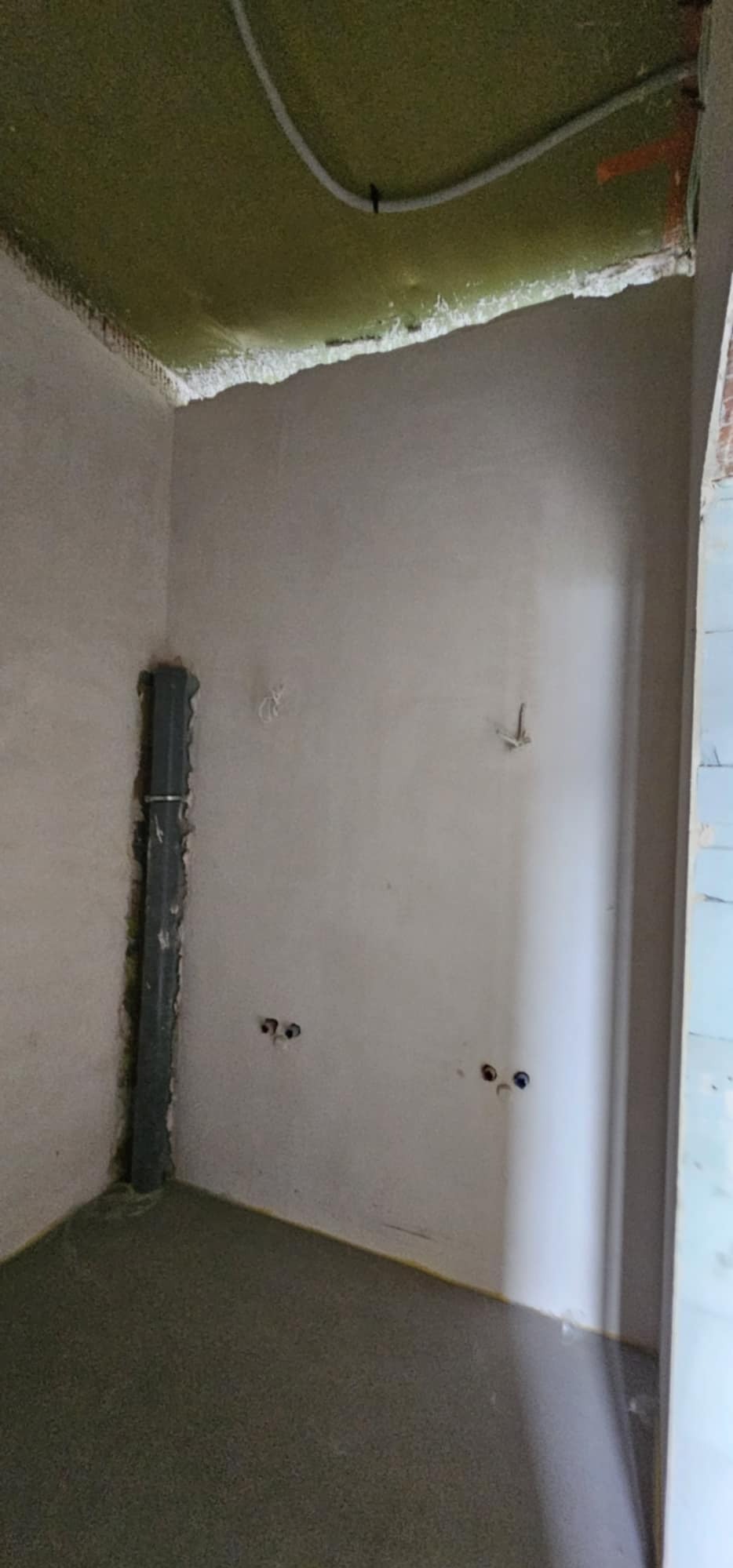 Surowe wnętrze w trakcie prac przygotowawczych: ściany pokryte szpachlą, widoczne puszki elektryczne, rury i izolacja sufitu.