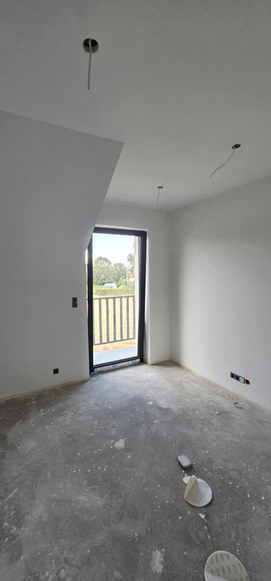 Surowe wnętrze z oknem balkonowym, świeżo pomalowane ściany na biało, widoczne wystające kable ze stropu i szpachlowana podłoga z widocznymi śladami prac wykończeniowych.