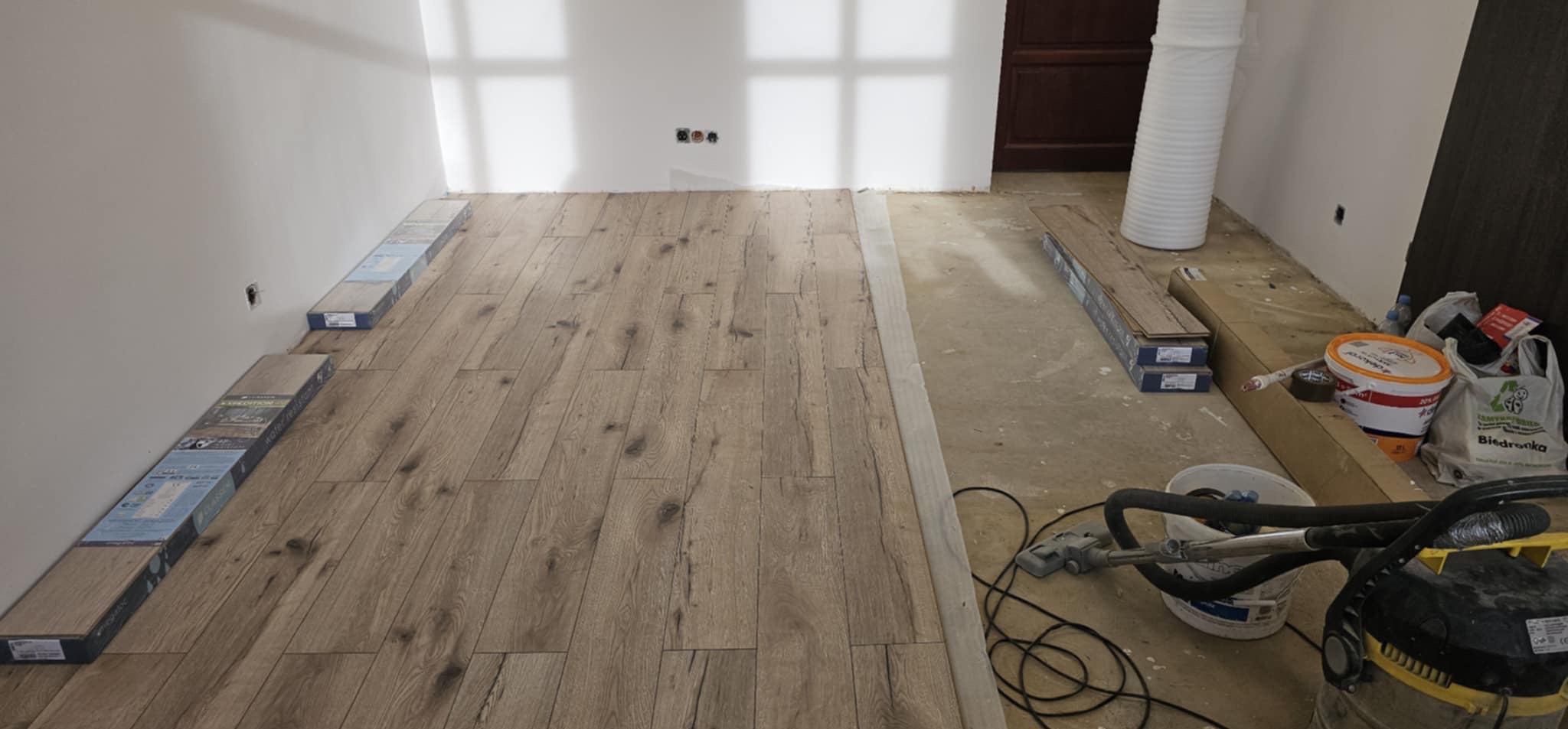 Układanie paneli podłogowych w trakcie prac, widoczne paczki paneli przy ścianach, częściowo ułożona podłoga oraz sprzęt do sprzątania podłogi.