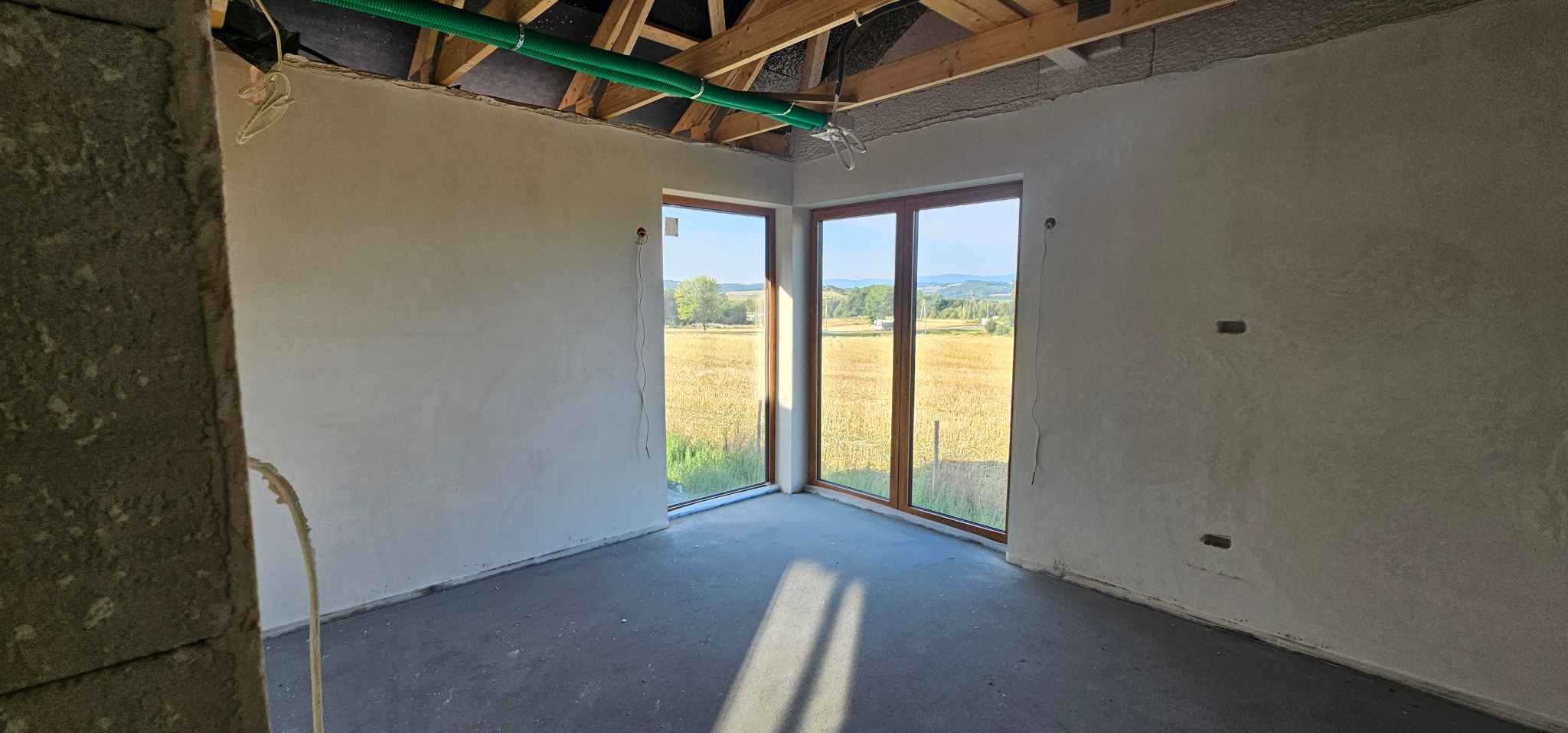 Wnętrze surowego domu po gładziowaniu ścian, z widokiem na pole przez okna. Widoczne instalacje pod sufitem i fragment ściany z surowego betonu po lewej.