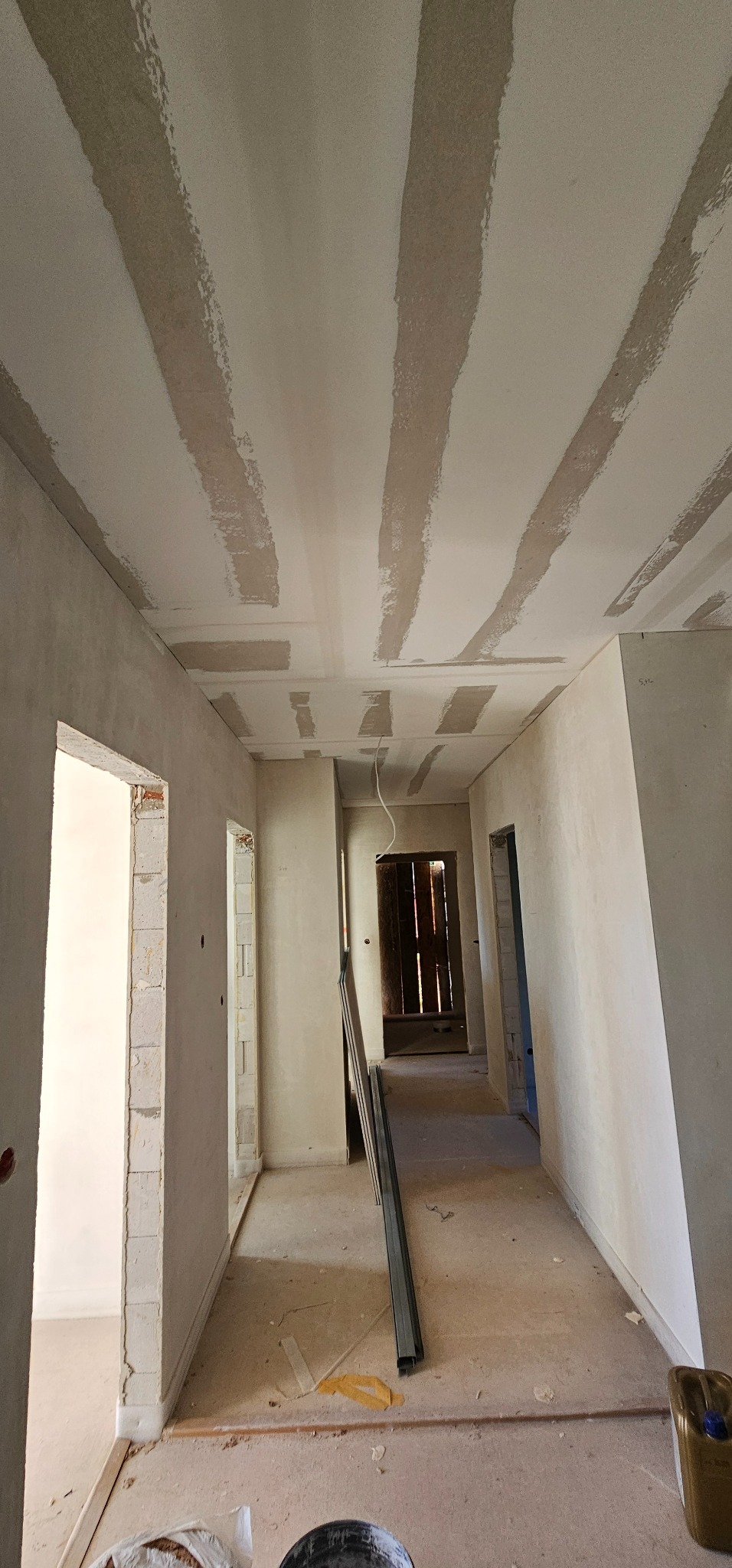 Wnętrze w trakcie prac wykończeniowych: sufit z płyt gipsowo-kartonowych ze śladami szpachlowania, widoczne profile i surowe ściany. Perspektywa korytarza.