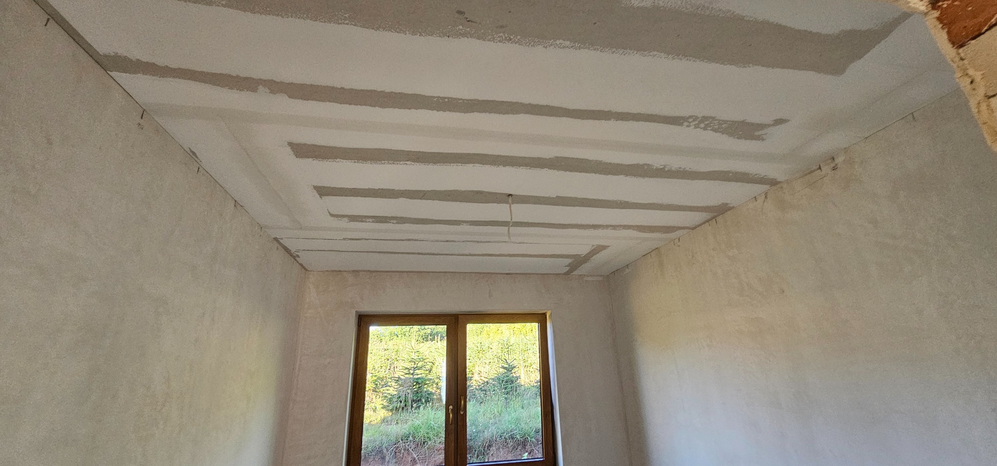 Wnętrze pokoju w trakcie prac wykończeniowych: surowa ściana, okno z widokiem na zieleń i sufit z widocznymi pasami gładzi w dekoracyjnym układzie.