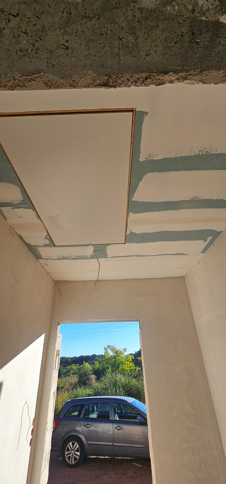 Montaż sufitu podwieszanego z płyty karton-gips z widoczną konstrukcją nośną i szpachlowanymi łączeniami, w tle widok na zewnątrz przez otwór okienny.