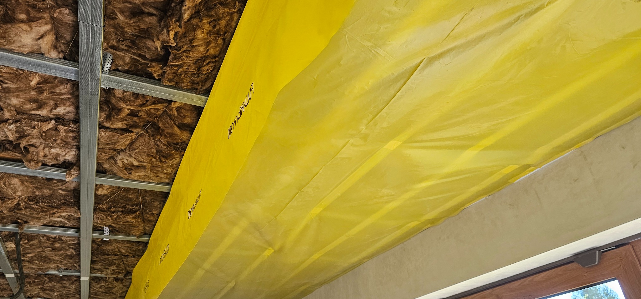 Izolacja poddasza wełną mineralną z folią paroizolacyjną, widoczna metalowa konstrukcja sufitu podwieszanego, jasne oświetlenie.