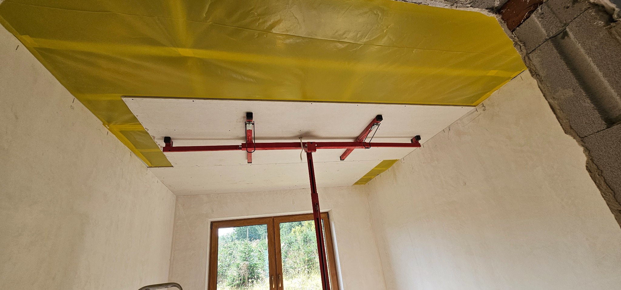 Montaż płyty kartonowo-gipsowej na suficie za pomocą podpór. Żółta folia zabezpieczająca. Widoczne okno i surowy fragment ściany.