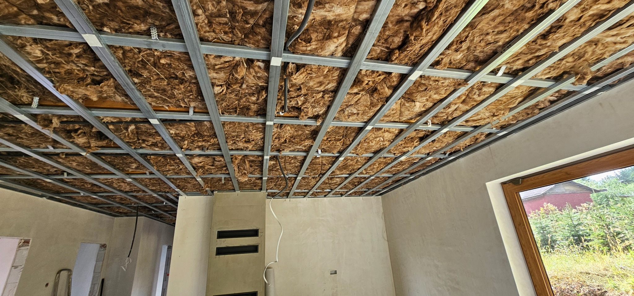 Sufit podwieszany z izolacją: metalowy stelaż wypełniony wełną mineralną. Widoczne fragmenty ścian w surowym stanie i okno z widokiem na zieleń.