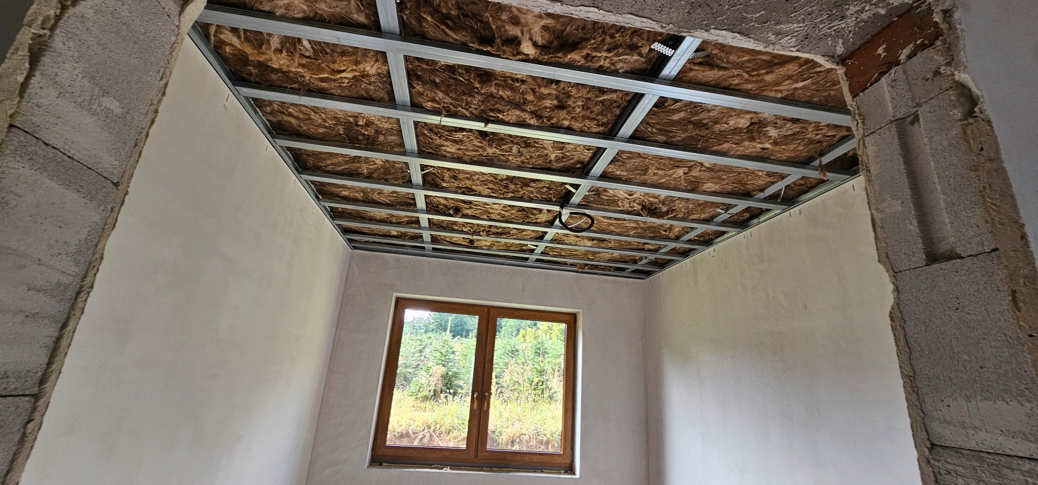 Wnętrze w trakcie prac: szkielet sufitu podwieszanego z izolacją, świeżo otynkowane ściany, okno z widokiem na zieleń. Surowy beton w narożnikach.