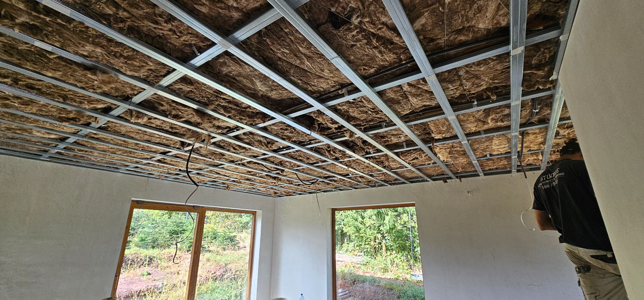 Sufit podwieszany z izolacją z wełny mineralnej, widok z dołu na metalową konstrukcję. W tle okno z widokiem na zieleń. Częściowo widoczna sylwetka pracownika.