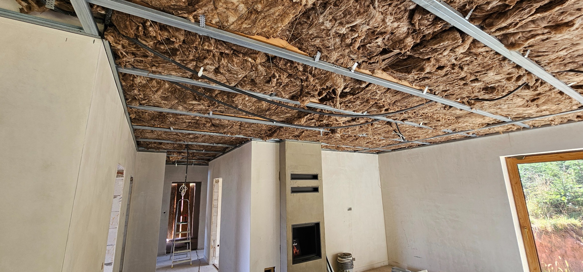 Sufit podwieszany z izolacją z wełny mineralnej i metalową konstrukcją w surowym wnętrzu domu. Widoczne przewody elektryczne i kominek.