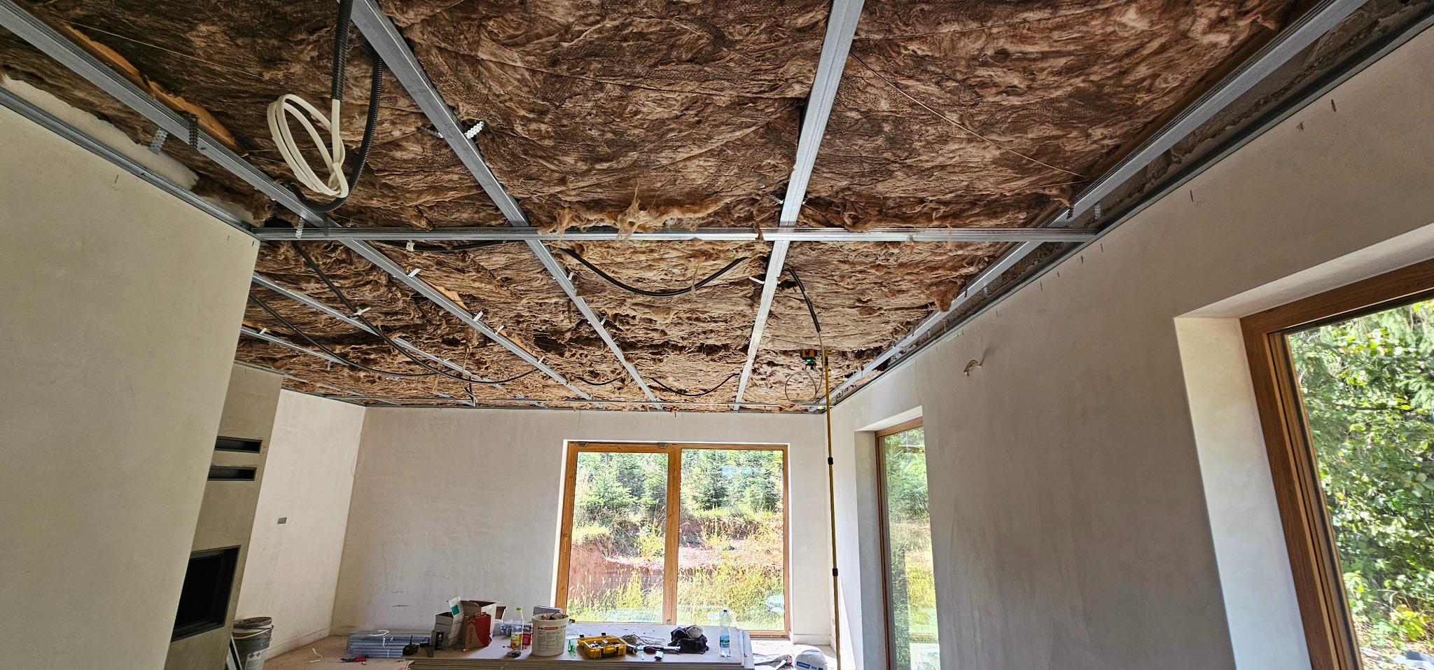 Wnętrze w trakcie prac: metalowa konstrukcja sufitu podwieszanego z ociepleniem z wełny mineralnej, ściany po gładziowaniu, okna z widokiem na zieleń.