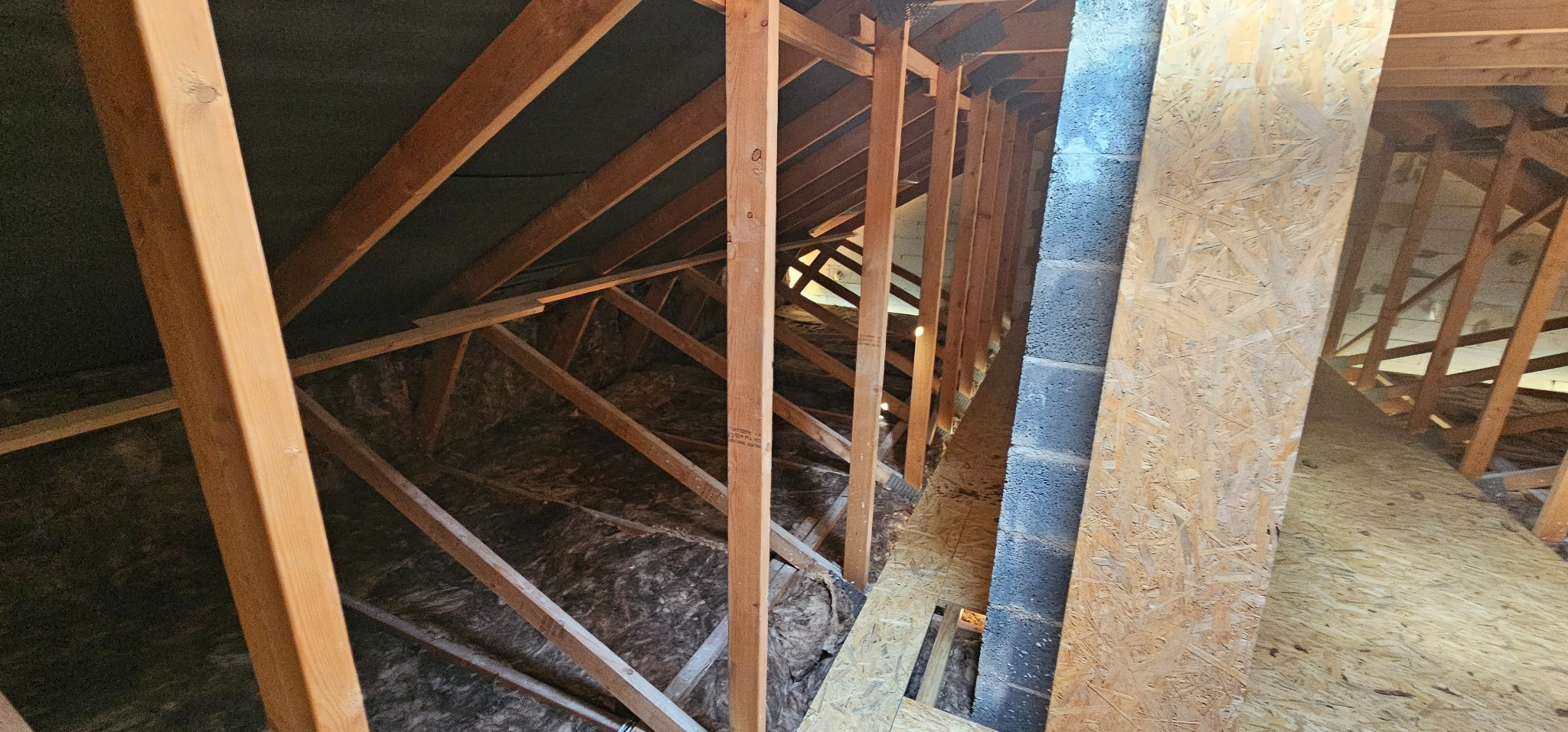 Skośny strych z drewnianą konstrukcją i izolacją z wełny mineralnej. Widoczne elementy więźby dachowej i komin z bloczków betonowych. Perspektywa z dołu.