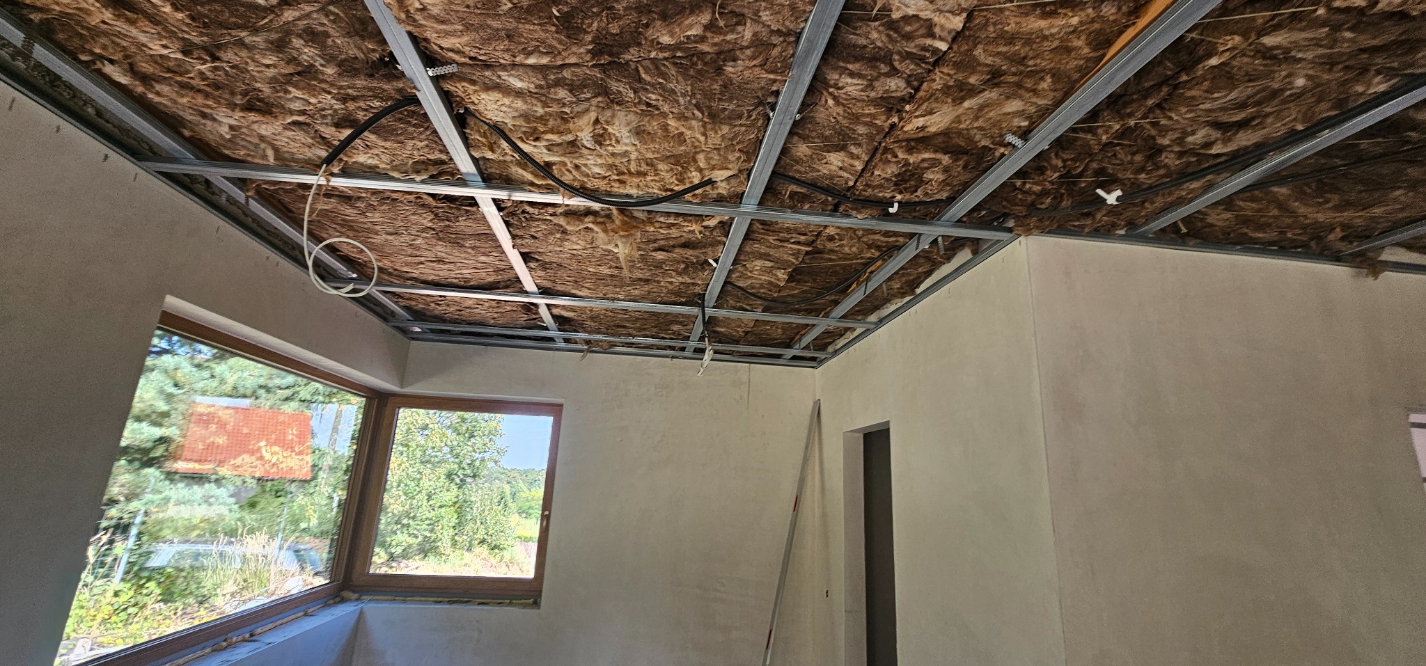 Montaż sufitu podwieszanego: metalowa konstrukcja z izolacją z wełny mineralnej, widok na okno i wstępnie wykończone ściany w surowym wnętrzu.