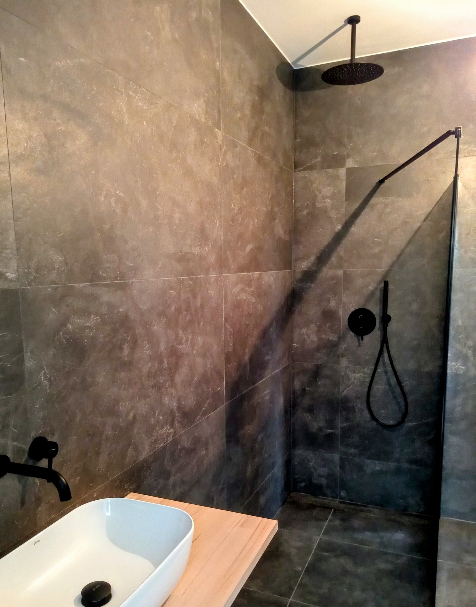 Nowoczesna łazienka z szarymi płytkami imitującymi kamień, czarną armaturą i białą umywalką na drewnianym blacie, widoczny deszczownica i bateria prysznicowa.