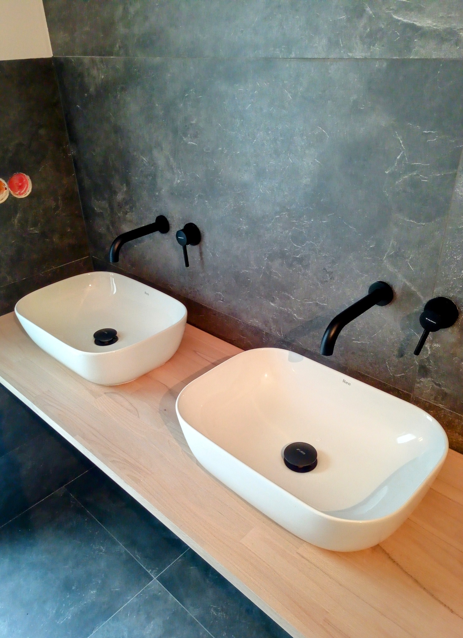 Dwie białe, prostokątne umywalki nablatowe z czarnymi bateriami na drewnianym blacie w łazience w Wągrowcu, z szarą, kamienną ścianą w tle i widocznymi puszkami instalacyjnymi.