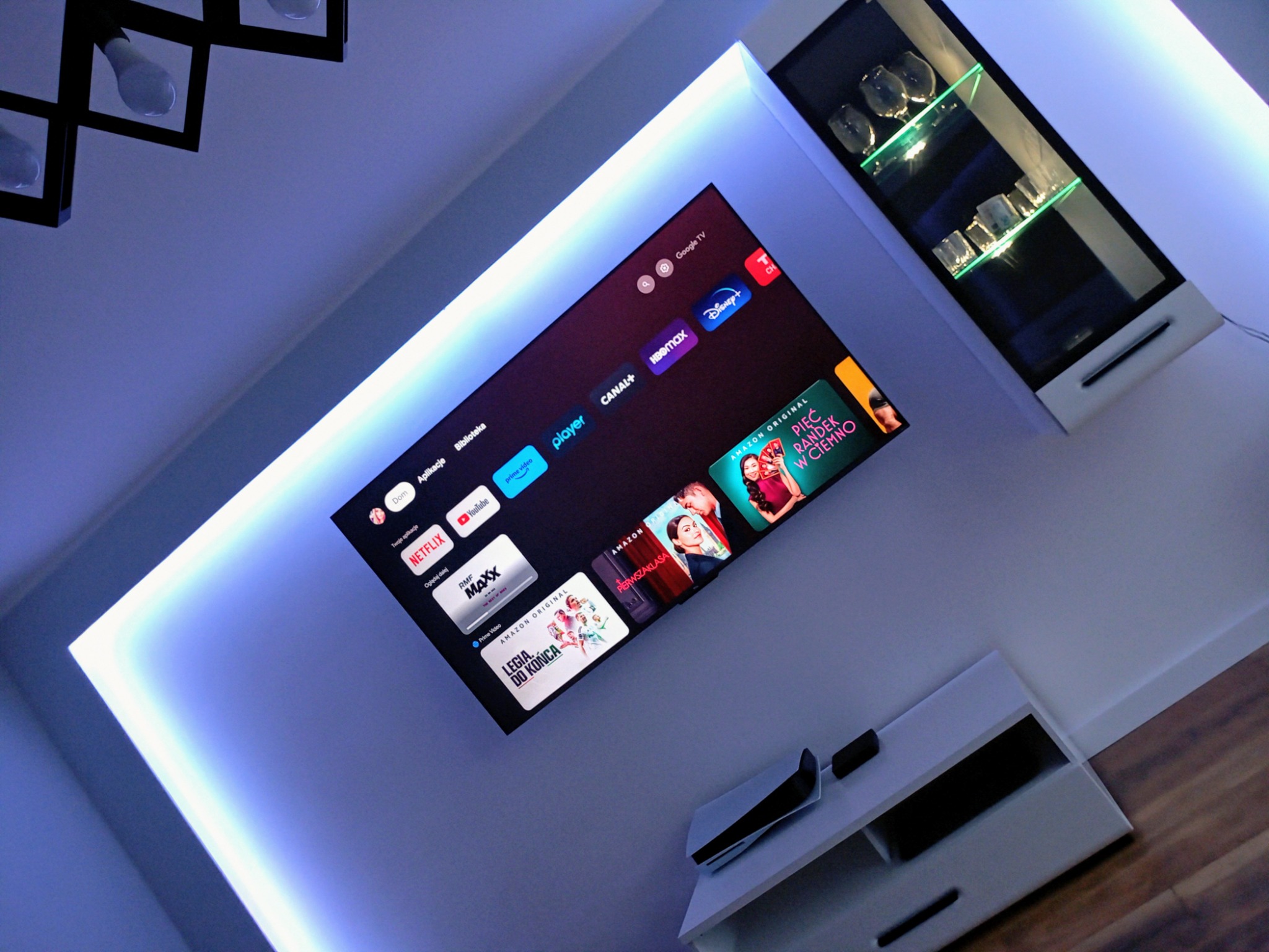 Nowoczesny salon z podświetlanym sufitem i ścianą, telewizorem Smart TV z menu aplikacji streamingowych, szklaną witryną z dekoracyjnym oświetleniem i drewnianą podłogą.