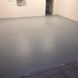 Szara podłoga w garażu pokryta żywicą z drobnymi, ciemnymi plamkami, widok na białe ściany i wejście do innego pomieszczenia.