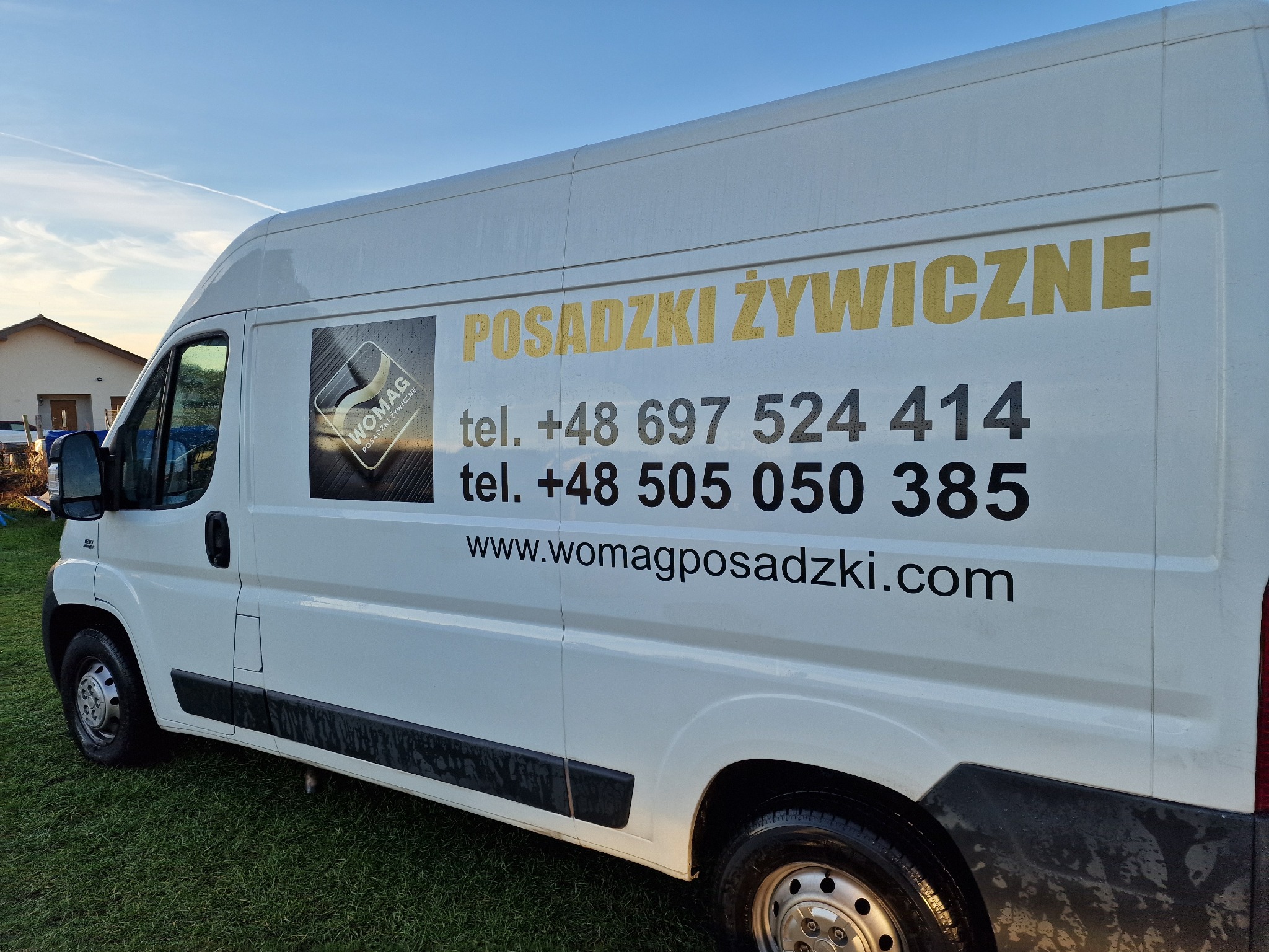 Biały samochód dostawczy firmy Womag Posadzki Żywiczne z numerami telefonów i adresem strony internetowej, zaparkowany na trawie.