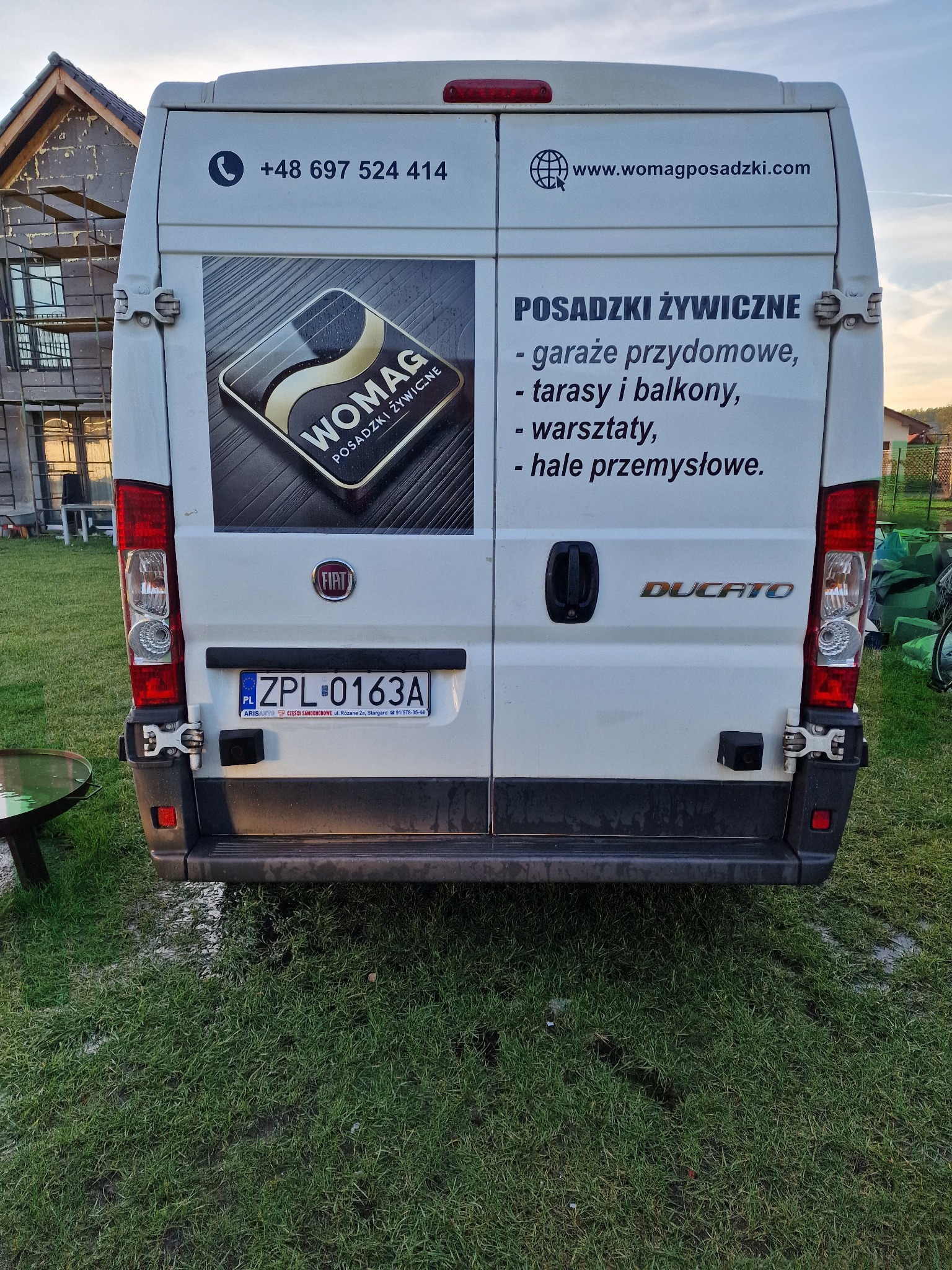 Tył białego samochodu dostawczego Fiat Ducato z reklamą firmy Womag, specjalizującej się w posadzkach żywicznych, zaparkowanego na trawniku przed budową.